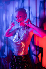 provocative woman in metallic top biting silver chain and looking at camera in blue and pink light near colorful graffiti.