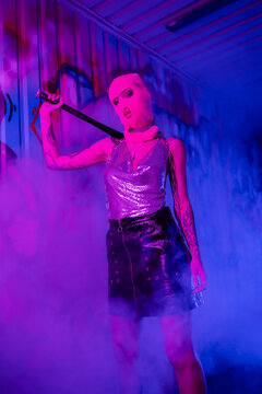 sexy woman in pink balaclava and silver top with leather skirt holding baseball bat near wall with graffiti in purple fog.