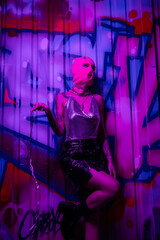 Fototapeta na wymiar provocative woman in pink balaclava and shiny top standing with silver chain near colorful graffiti in purple neon light.
