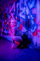 Fototapeta na wymiar full length of woman in stylish clothes and balaclava sitting near wall with graffiti in blue and purple lighting.