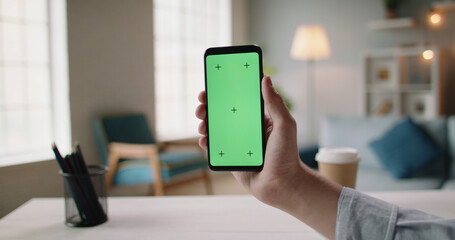 Close up shot of hand of guy holding a smartphone in cozy living room. A green screen mock up chroma key template 