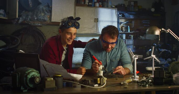 Portrait of Teenage Boy and his Father in Workshop Working Together on Soldering Wires and Circuit Boards for School Science Fair. Man and Son Bonding, Sharing their Hobby, Celebrating their Success