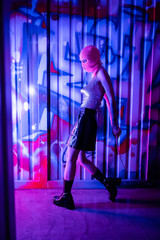 Obraz na płótnie Canvas side view of sexy woman in stylish outfit and balaclava standing with chain near wall with graffiti in blue and purple light.