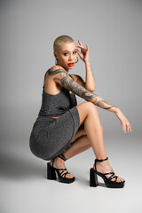 full length of fashionable tattooed woman on heels sitting on haunches and looking at camera on grey background. © LIGHTFIELD STUDIOS