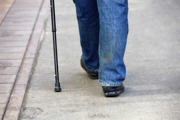 Man in jeans walking with cane on a street, legs on sidewalk. Concept of disability, limping senior adult, diseases of the spine