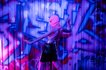 side view of provocative woman in black leather skirt and balaclava posing with chain near colorful graffiti in blue and purple light.