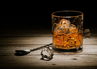 Glass of single malt whiskey with bar spoon and ice cubes on wooden background.