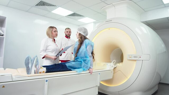 Woman doctor radiologist explains good results of CT scanning for young female patient, showing the snapshot with images, observing and analyzing CT scan in modern clinic beside modern CT scanner