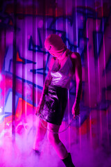 sexy woman in balaclava and shiny top with leather skirt standing with chain near colorful graffiti in purple smoke.