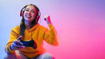 A young Caucasian beautiful woman in a yellow hoodie playing a video game using a joystick and headphones with fun, focus, surprise, and a winning victory face with blue and red neon light. © artitwpd