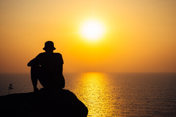 Fototapeta na wymiar The silhouette of a man in a cap against the background of a bright sunset and the endless sea. A thoughtful adventurer, thoughts about the future and the meaning of life