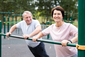 Aged man and woman doing exercises on sports bars in open-air sports area