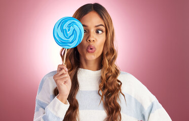 Portrait, funny face and lollipop with a woman on a pink background in studio holding giant candy. Comic, comedy and sweet with a playful young female feeling silly while eating a blue sugar treat
