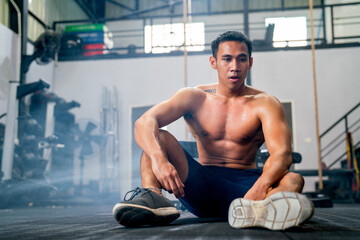 Front view of Asian sport man with shirtless sit on floor of fitness gym after finish of exercise.
