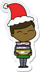 sticker cartoon of a smiling boy with stack of books wearing santa hat