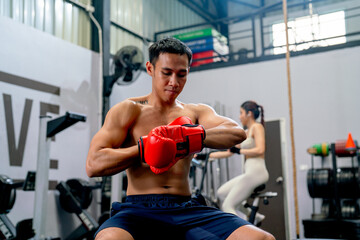 Fototapeta na wymiar Asian sport man sit on chair and try to wear boxing glove in front of the woman exercise in the back of fitness gym.