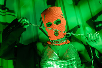provocative woman in knitted balaclava and silver top holding metallic chain near wall with graffiti in green lighting.