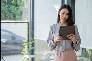 Asian woman relaxing hands using tablet surfing internet writing diary smiling cheerfully while sitting in cafe near large window