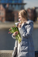 Fototapeta premium A young blonde woman in a coat stands with a bouquet of red tulips on the embankment in the park. The concept of the spring holiday is March 8, Easter, Women's Day.
