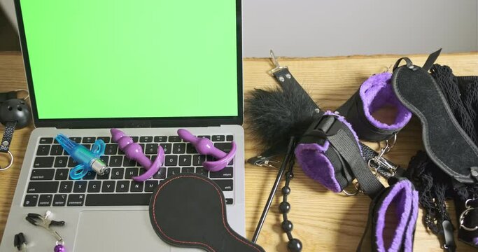Set of sex toys lay next to laptop with green screen. Anal stimulators and vibrator lie on laptop keyboard. Concept of an online sex dating service, a bdsm site, a sex shop.