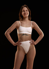Fototapeta premium Portrait of positive, smiling , attractive young girl with tattoo and piercing posing in white cotton underwear against black studio background. Body and skin care, figure, fitness, wellness concept