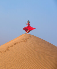 Beautiful young woman with a red dress dancing and posing on the dunes in the desert. - 584307403