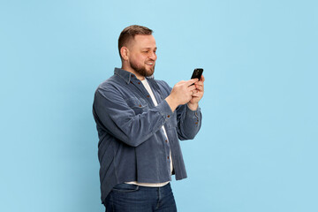 Happy bearded man in jeans jacket looking at cell phone and smiling over blue background