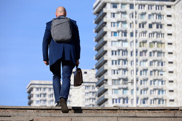Businessman with backpack and leather briefcase in hand climbing stone stairs in city. Concept of career, success, moving to the top