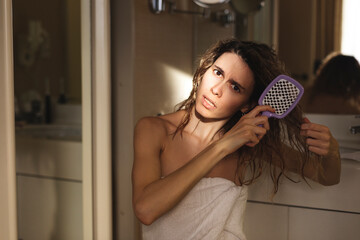 Close up of sad young woman in white towel brush her damaged and tangled wet hair. Morning routine....