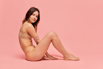 Obraz na płótnie Canvas Portrait of young, beautiful, slim girl in beige underwear posing against pink studio background. Self-care and acceptance. Concept of body and skin care, figure, fitness, health, wellness.