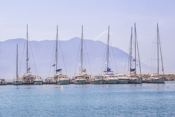 Fototapeta na wymiar Several yachts moored in one row in a light morning haze against the backdrop of a mountain in the port of Budva, Montenegro. Gentle seascape with single-masted passenger boats in the Adriatic Sea