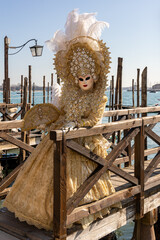 Venice carnival 2023, an unrecognizable masked model posed near a canal in Venice. A vintage costume with blue skys and sea to the background .