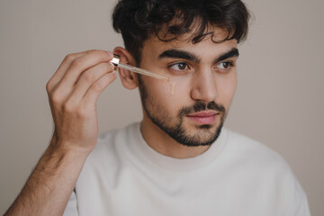 Close up portrait of a young man applying face body oil, moisturizing serum isolated on gray...