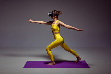 Fototapeta premium Young slim girl in yellow sportswear training, doing yoga exercises in VR glasses against purple background. Flexibility and strength. Concept of body , figure, fitness, health, wellness.