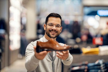 A happy young smart casual man is standing at shoe store and showing leather shoes at the camera.