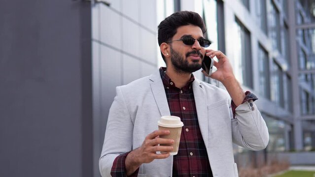 Energetic Indian mixed race businessman talking on cellular device while wearing sunglasses, holding a cup of coffee, dressed in a stylish and cool outfit outdoors. Business man speaking on mobile.