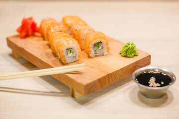 Traditional Japanese food. Freshly made sushi rolls of fish, rice, cheese, cucumber and sesame seeds served on a wooden board. Next to soy sauce, ginger, wasabi, chopsticks.