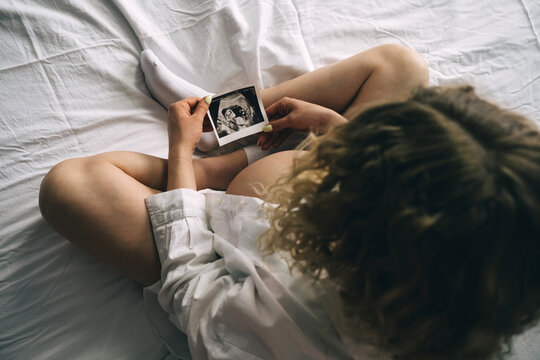 The pregnant girl holding an ultrasound picture in her hands while sitting on the bed