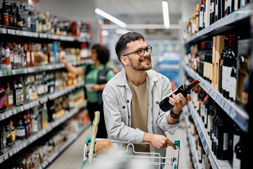 Happy man choosing wine bottle from shelf while shopping in supermarket.