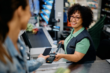 Happy cashier holds credit card reader while customer is paying with smart phone at supermarket. - obrazy, fototapety, plakaty