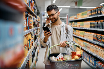 Young man using cell phone while shopping in supermarket. - obrazy, fototapety, plakaty