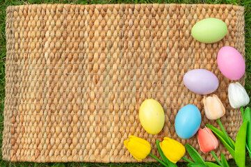 Happy Easter holiday greeting card concept. Colorful Easter Eggs and spring flowers on Papyrus rattan weave texture background. Flat lay, top view, copy space.