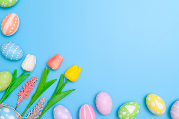 Happy Easter holiday greeting card design concept. Colorful Easter Eggs and spring flowers on blue background. Flat lay, top view, copy space.