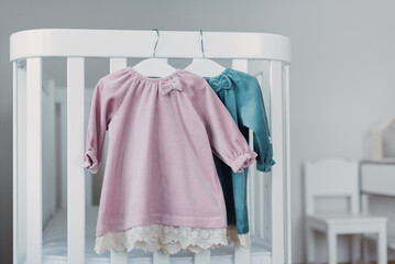 Pink, blue dresses for baby girl on white hanger near white bed in nursery. Set of baby clothes and...