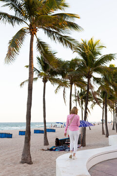 Selective focus back view of slim young blond woman in pink shirt and white pants walking on low wall along the beach, Fort Lauderdale, Florida, USA