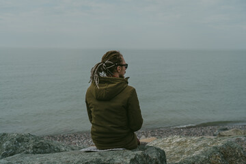 Young man with dreadlocks sits in lotus position on stone beach near sea water and meditates, enjoying peace and tranquility of nature.