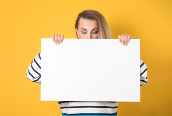 Close-up woman behind white board and looking down to empty space, isolated on yellow background. Pretty girl showing blank space for text near her head