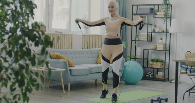 Portrait of slender elderly lady in activewear exercising with resistance band at home. Healthy senior people and retirement concept.