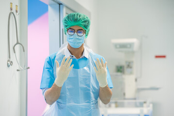 Fototapeta premium Man male obstetrician doctor portrait in uniform coat gown with white hand grove. Medical senior gynecologist stand by give birth wear blue gown coat stand in delivery room pregnant hospital.