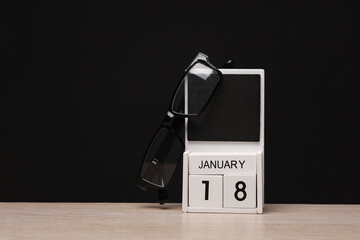 White wooden block monthly calendar with the date january 18 and eyeglasses on the table, black blackbackground. Planning